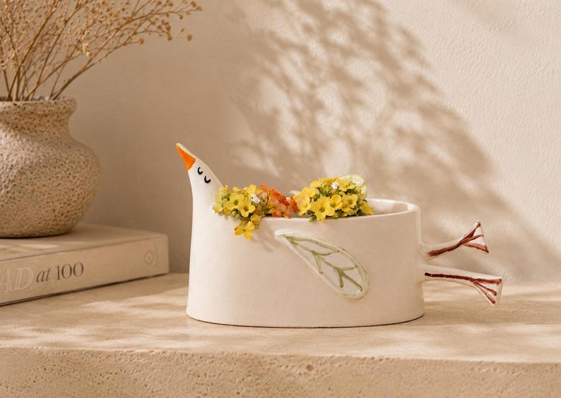 A white ceramic bird planter filled with yellow flowers, resting on a beige surface with a book and vase in the background.