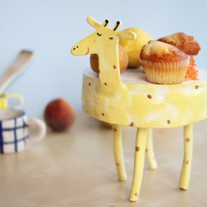 Puede incluir: Un caprichoso soporte para pasteles con forma de jirafa amarilla con lunares marrones, que contiene magdalenas y fruta. La jirafa tiene un cuello largo y una cara sonriente. Otras tazas de cerámica y un melocotón están en el fondo.