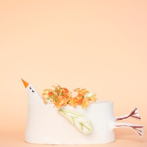 The white ceramic bird planter filled with orange flowers displayed against a solid peach-colored background.