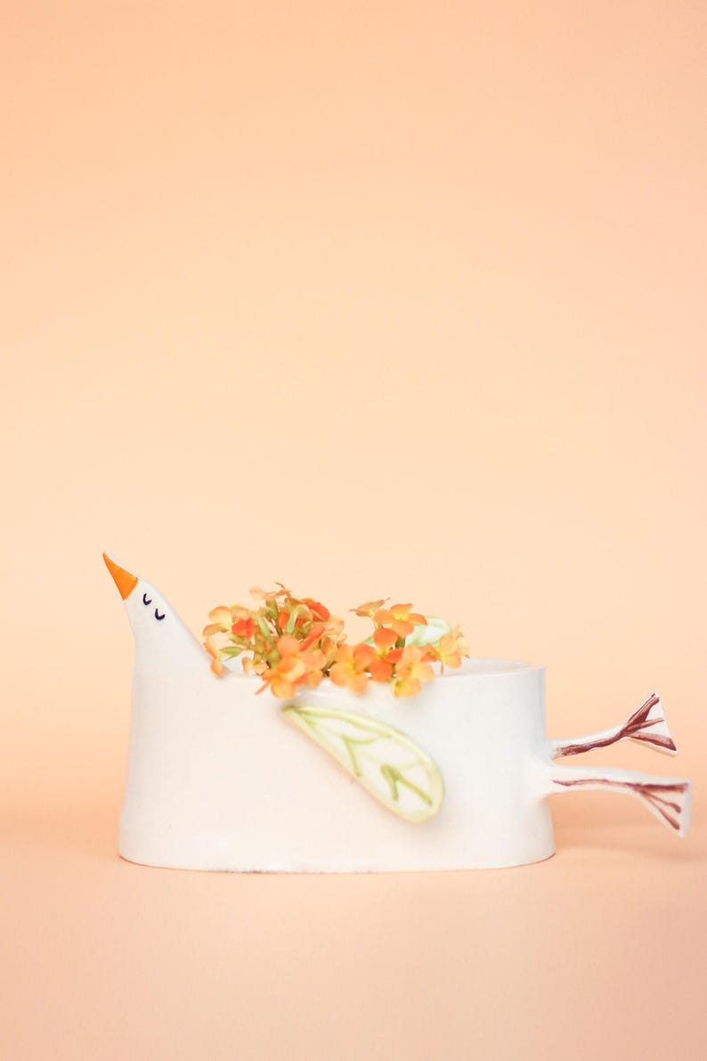 The white ceramic bird planter filled with orange flowers displayed against a solid peach-colored background.