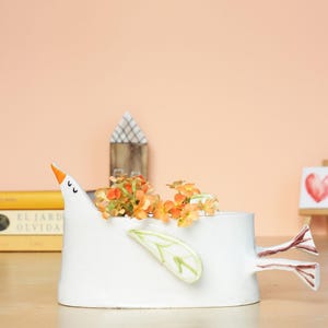 The white ceramic bird planter with orange flowers placed in front of a warm peach-colored wall and stacked books.