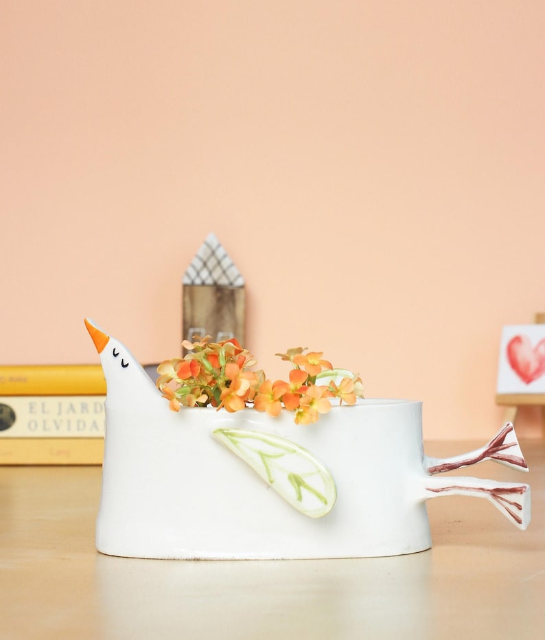 The white ceramic bird planter with orange flowers placed in front of a warm peach-colored wall and stacked books.