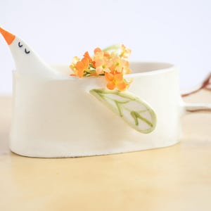 A close-up side profile of the white ceramic bird planter holding orange flowers against a clean white background.