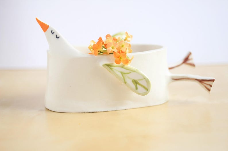 A close-up side profile of the white ceramic bird planter holding orange flowers against a clean white background.