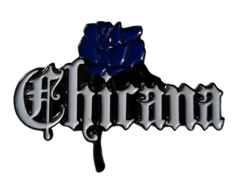 May include: A white enamel pin with the word "Chicana" in black script with a blue rose on top.