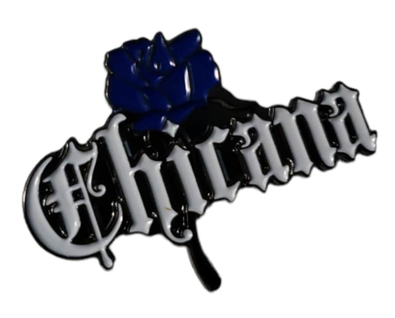 May include: A black and white enamel pin with the word "Chicana" in a stylised font. A blue rose is attached to the left side of the word.