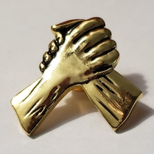 May include: Gold-toned metal pin featuring two hands clasped together in a handshake. The pin is a symbol of unity and friendship.