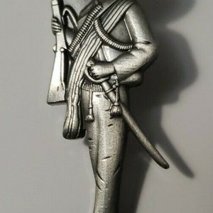 Op de afbeelding: Een zilverkleurig metalen beeldje van een man met een sombrero die een geweer en een zwaard vasthoudt. Het beeldje staat op een witte achtergrond.