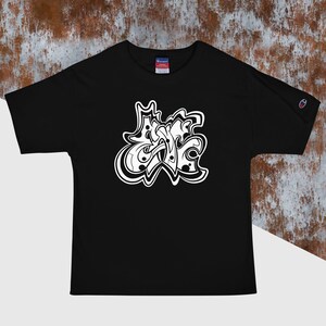 以下が含まれることがあります： 黒いチャンピオンTシャツ。前面には白いグラフィティ風のデザインが施されています。このデザインは、大胆で芸術的な美しさを持つ、線と形の抽象的な配置です。Tシャツは半袖で、クルーネックです。