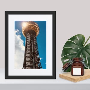 Puede incluir: Impresión enmarcada de la Sunsphere, una torre dorada con cúpula y estructura de celosía, sobre un cielo azul brillante con nubes. La imagen está en un marco negro, con un pequeño tarro y una planta en primer plano.