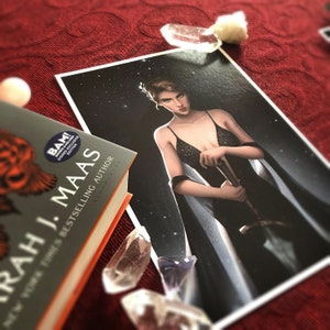 May include: A book by Sarah J. Maas with a signed edition sticker, next to a print of a woman in a black dress holding a sword. Clear quartz crystals and a white sphere are scattered around the book and print on a red surface.