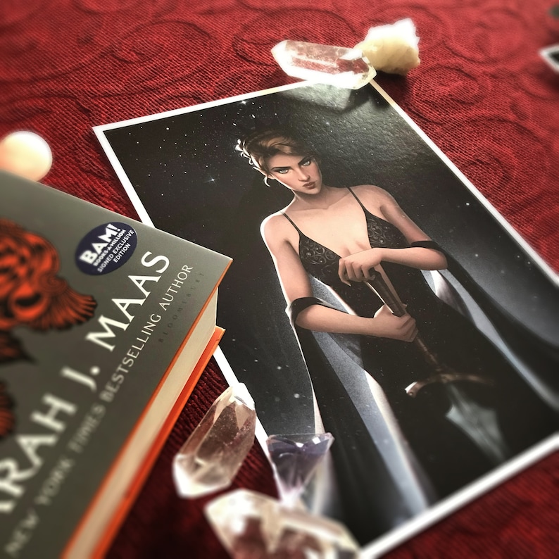 May include: A book by Sarah J. Maas with a signed edition sticker, next to a print of a woman in a black dress holding a sword. Clear quartz crystals and a white sphere are scattered around the book and print on a red surface.
