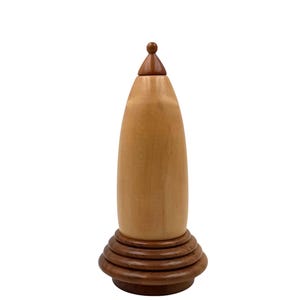 May include: A handcrafted wooden urn with a light brown body and a dark brown base. The urn has a conical shape with a small, rounded lid. The base features concentric rings. The urn is made of wood.