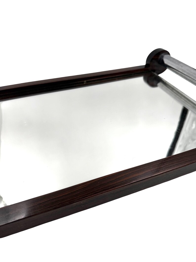 Puede incluir: Una bandeja rectangular con superficie de espejo y marco de madera marr&oacute;n oscuro. La bandeja tiene un asa cromada y est&aacute; inclinada hacia el espectador, mostrando sus cualidades reflectantes.