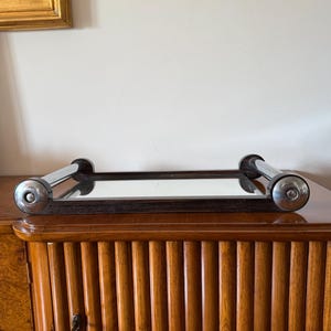Puede incluir: Una bandeja rectangular con espejo, borde de madera oscura y asas cromadas. La bandeja se encuentra sobre un mueble de madera con detalles verticales acanalados. El mueble es de color marr&oacute;n medio c&aacute;lido.