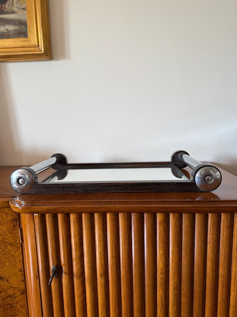 Puede incluir: Una bandeja rectangular con espejo, borde de madera oscura y asas cromadas. La bandeja se encuentra sobre un mueble de madera con detalles verticales acanalados. El mueble es de color marr&oacute;n medio c&aacute;lido.