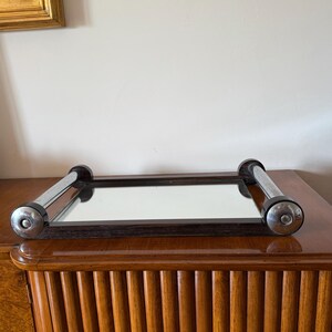 Puede incluir: Un espejo rectangular con marco de madera oscura y asas cromadas. El espejo est&aacute; colocado sobre un mueble de madera con un dise&ntilde;o acanalado. Las asas cromadas tienen detalles negros en los extremos. El espejo refleja la habitaci&oacute;n.