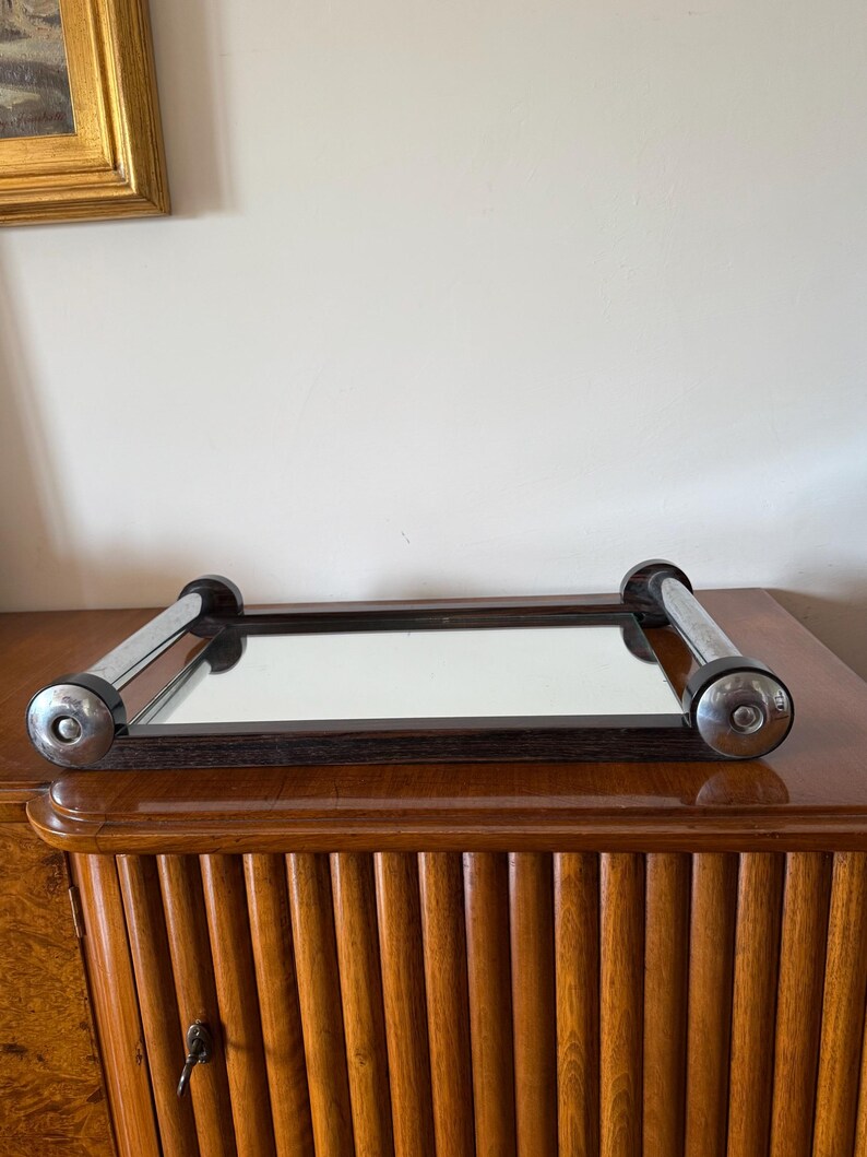 Puede incluir: Un espejo rectangular con marco de madera oscura y asas cromadas. El espejo est&aacute; colocado sobre un mueble de madera con un dise&ntilde;o acanalado. Las asas cromadas tienen detalles negros en los extremos. El espejo refleja la habitaci&oacute;n.