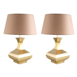 May include: A pair of table lamps. Each lamp has a beige lampshade and a gold-coloured base with a geometric design. The base has a square bottom and an hourglass-like shape. The lamps are set against a white background.