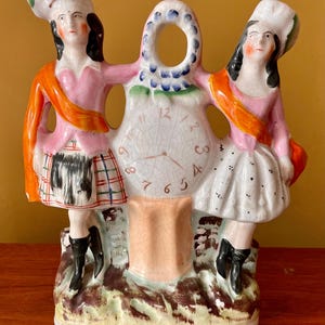 Puede incluir: Figura de cerámica con dos figuras vestidas con trajes escoceses a ambos lados de una esfera de reloj. Las figuras visten chaquetas rosas, fajas naranjas y faldas escocesas. El reloj tiene números romanos y está rodeado de un diseño floral.
