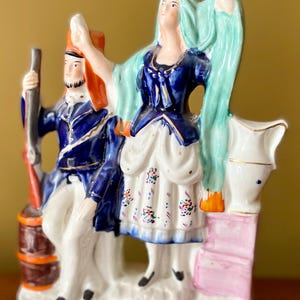 Puede incluir: Figura de cerámica que representa a un hombre con uniforme azul y a una mujer con vestido azul, con el texto "HIGHLAND JESSIE" en la base. La mujer tiene el pelo largo y verde. La figura está pintada con colores brillantes.
