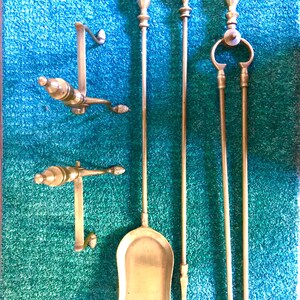 Peut inclure: Un ensemble d'outils de cheminée en laiton antique, comprenant une pelle, un tisonnier, des pinces et deux supports muraux. Les outils ont des poignées ornées et sont disposés sur une surface texturée turquoise.
