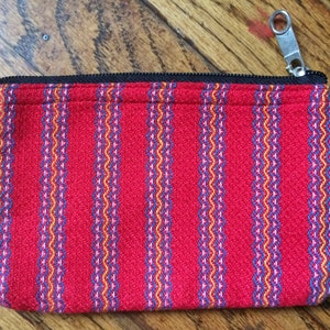 May include: Red zippered pouch with a striped pattern in blue, yellow, and white. The pouch is made of a woven fabric.
