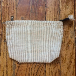 May include: A small, beige zippered pouch with a white and brown striped pattern. The pouch has a black zipper pull and a black strap.