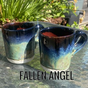 May include: Two ceramic mugs with a distinctive glaze. The mugs display a gradient of colours, including blue, green, and brown, with a white base. The text "FALLEN ANGEL" is at the bottom. These mugs are suitable for coffee or tea.