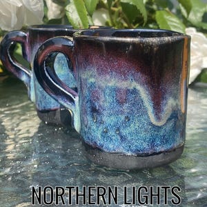 Puede incluir: Dos tazas de cerámica con un diseño de Auroras Boreales. Las tazas tienen una base oscura, que transiciona a tonos azules y morados, con un acabado brillante. El texto "NORTHERN LIGHTS" está en la parte inferior de la imagen.