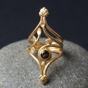 May include: A gold-colored ring with an ornate design featuring a central brown gemstone. The ring has a unique shape with leaf-like accents and a decorative top. The ring is displayed on a gray stone against a dark background.