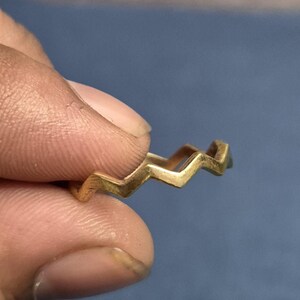Peut inclure: Une bague dorée avec un motif en zigzag. La bague est tenue entre les doigts sur un fond bleu. Le design de la bague est une série de pics et de creux angulaires reliés.