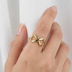 May include: A polished gold-tone ring featuring a continuous infinity symbol design. The band is smooth and reflective, forming a horizontal figure-eight shape that sits across the finger. The metal has a bright, warm yellow metallic finish.