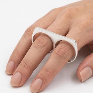 Peut inclure: Une bague argentée à deux doigts avec une barre rectangulaire reliant deux anneaux circulaires. La bague est portée sur deux doigts, mettant en valeur son design moderne. La bague est en métal brillant.