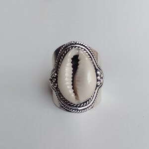 May include: A silver ring with a cream-coloured cowrie shell centrepiece. The ring has a wide band with decorative rope and dot detailing around the shell. The shell is oval-shaped and has a natural, textured appearance.