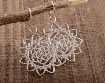 May include: A pair of silver dangle earrings featuring a circular, openwork design. The center of each earring displays a geometric pattern of interlocking circles, surrounded by a border of small, heart-shaped cutouts. The earrings hang from silver hooks.