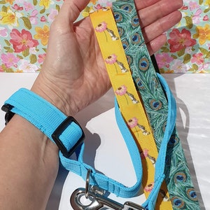 May include: A light blue dog leash and matching wrist strap with a black buckle. Two additional patterned ribbons are included: one yellow with pink flowers and one green with peacock feathers. A silver metal clasp is attached.
