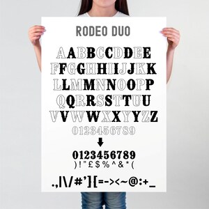 May include: A white poster with the text "RODEO DUO" in gray at the top. The poster features a black and white alphabet and numbers in a western-style font. The poster also includes punctuation marks and symbols.
