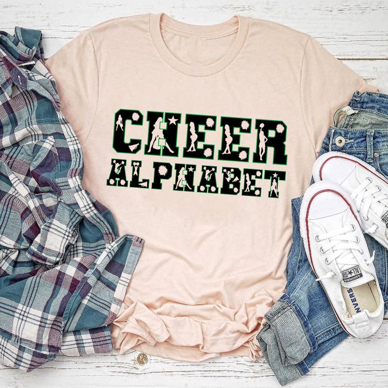 Puede incluir: Una camiseta rosa con el texto "CHEER ALPHABET" en letras negras con contorno verde. Las letras est&aacute;n formadas por animadoras en varias poses.