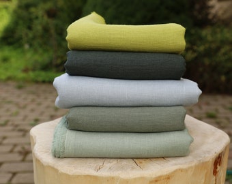 May include: Stack of five fabric swatches in various shades of green, including a light green, a dark green, a gray-green, a light blue-green, and a dark green. The swatches are folded and stacked on top of each other on a wooden surface.