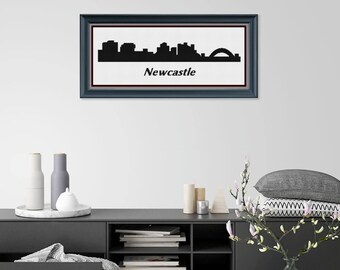 May include: A framed black and white silhouette of the Newcastle skyline, including the Tyne Bridge, against a white background. The word "Newcastle" is printed below the skyline. The frame is dark blue with a maroon inner border. The artwork is displayed above a black cabinet.