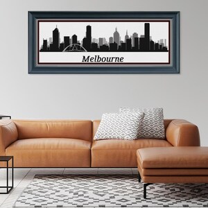 May include: A framed black and white silhouette of the Melbourne skyline, with the city's name in white text below. The artwork is displayed above a brown leather sofa with decorative pillows. A matching ottoman and a black side table complete the living room setting.