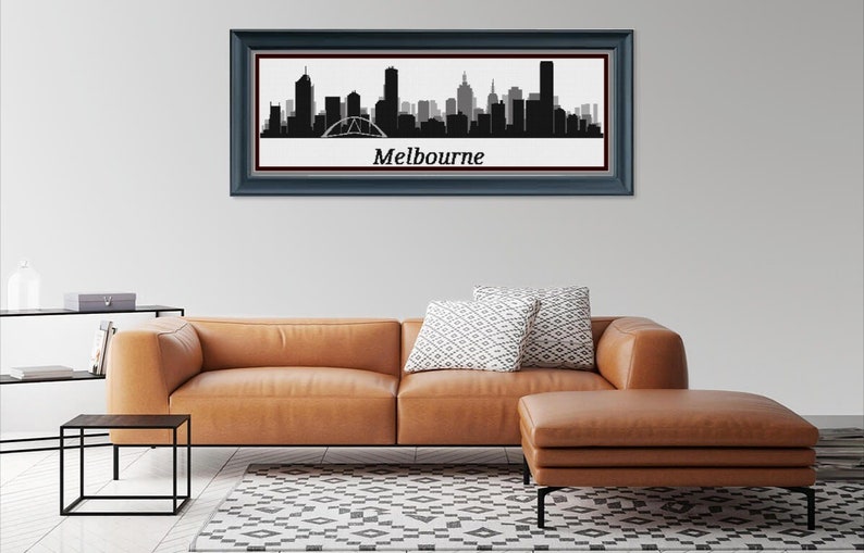 May include: A framed black and white silhouette of the Melbourne skyline, with the city's name in white text below. The artwork is displayed above a brown leather sofa with decorative pillows. A matching ottoman and a black side table complete the living room setting.
