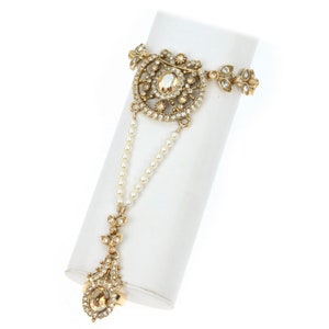 May include: A gold-tone hand and finger ring with a large, ornate, crystal-encrusted design. The ring is connected to a delicate pearl chain.
