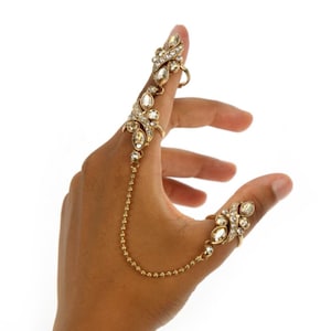May include: A gold ring set with clear rhinestones, featuring a connected chain that extends to a second ring on the thumb. The rings are designed with intricate floral patterns.