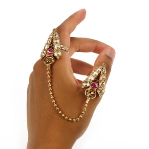 May include: A gold ring with a chain connecting two ornate, pointed sections. Each section is decorated with clear and pink rhinestones.