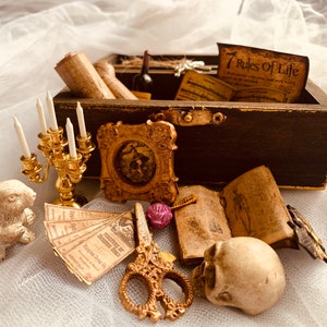 Könnte beinhalten: Eine Holzkiste mit einem goldenen Verschluss, gefüllt mit verschiedenen Gegenständen, darunter Kerzenleuchter, ein Buch, ein Schädel und eine Schriftrolle. Die Kiste ist geöffnet und die Gegenstände fallen heraus.