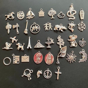 Könnte beinhalten: Eine Sammlung von 35 silbernen Anhänger-Anhängern mit verschiedenen Designs, darunter Tiere, Symbole und Wörter. Die Anhänger sind in Reihen auf einem schwarzen Hintergrund angeordnet.