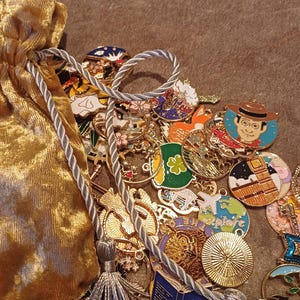 Könnte beinhalten: Ein goldfarbener Samtbeutel mit einem silbernen Kordelzug, gefüllt mit einer Vielzahl von bunten Emaille-Anhängern. Die Anhänger zeigen verschiedene Designs, darunter Tiere, Gebäude und abstrakte Formen.