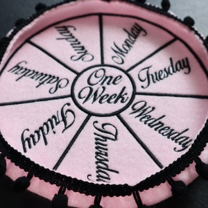 May include: A pink felt wheel with black trim and pom-poms around the edge. The wheel is divided into seven sections, each with a day of the week written in black embroidery. The center of the wheel says "One Week".